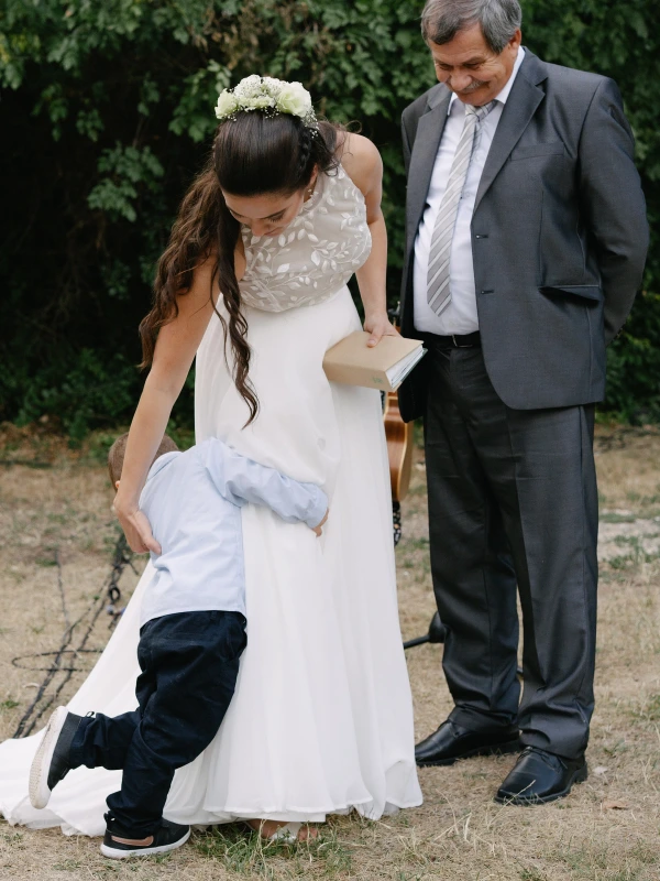 Bride in lace gown with father walking during ceremony, young boy holding her dress train outdoors.