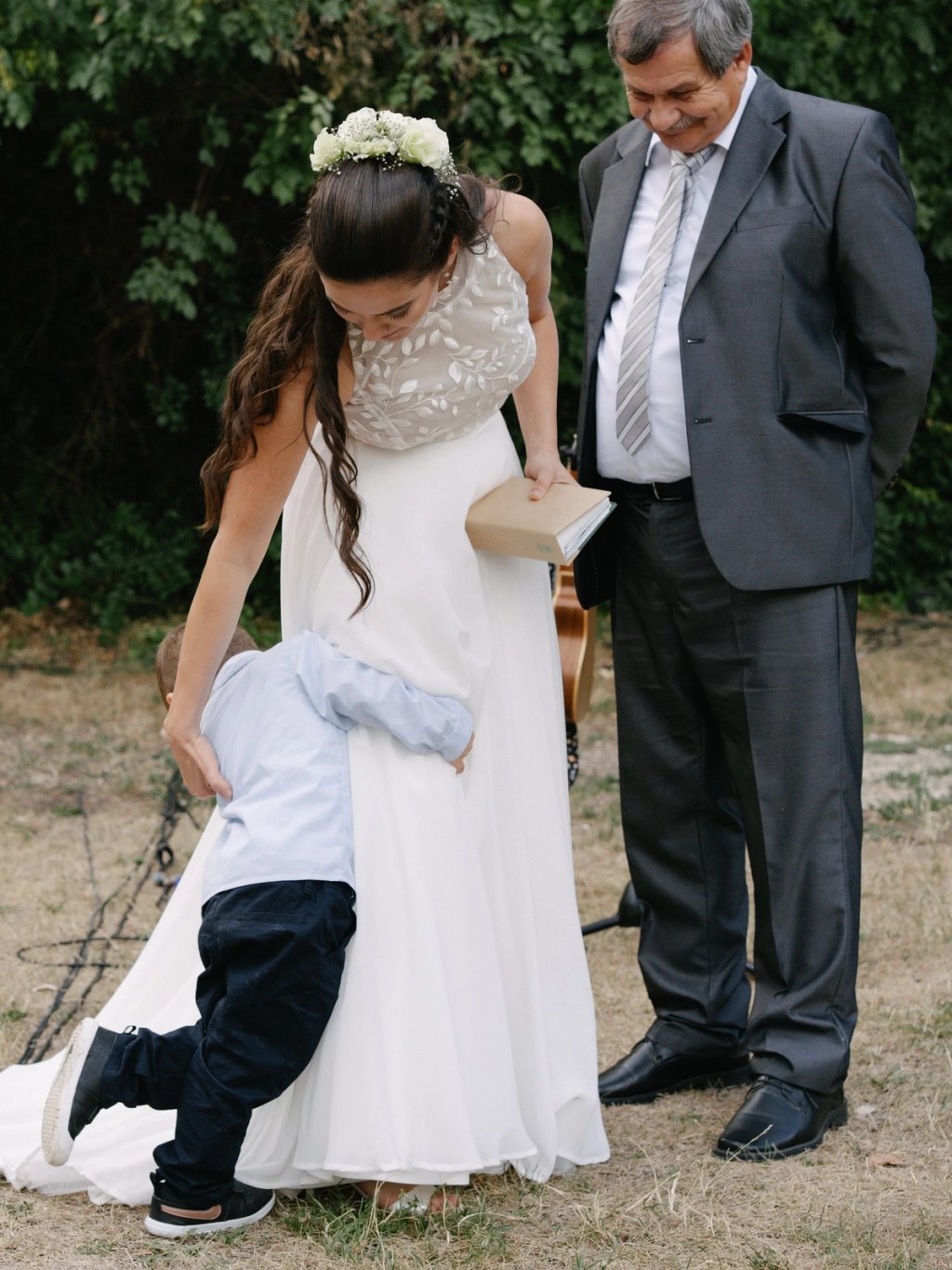 Bride in lace gown with father walking during ceremony, young boy holding her dress train outdoors.