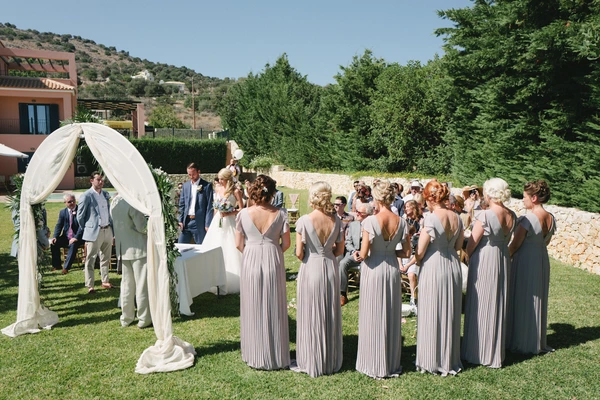Outdoor ceremony under white satin chuppah — bridesmaids in grey look up, green hills and terracotta house