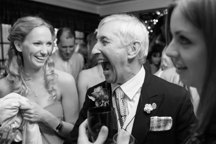 Elderly man laughing joyfully at wedding reception, holding drink, surrounded by smiling women in black and white photo.