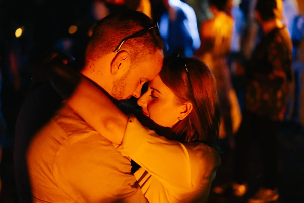 Bride and groom embrace warmly on dance floor — evening reception lighting, intimate moment, golden ambient glow.