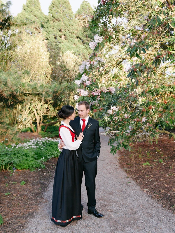The bride in a Norwegian bunad and the groom in a dark suit stand facing each other on a gravel path with a flowering tree overhead, sharing a quiet, affectionate moment. Documentary wedding portrait at the Royal Botanic Garden Edinburgh.