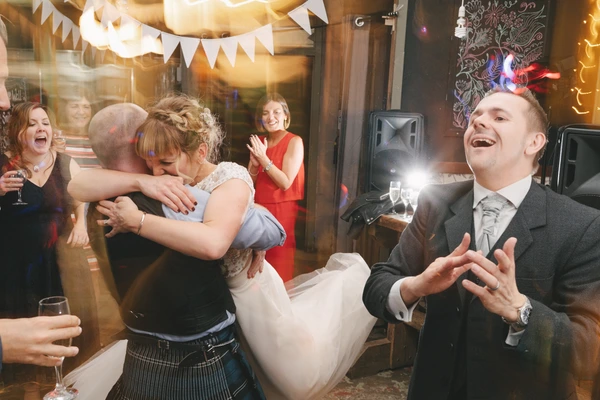 Groom watches the bride being lifted on a guest's shoulder — celebrating guests, paper garland