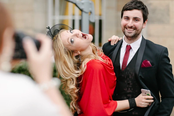 A woman in a red dress and a man in a tuxedo pose for a photograph, laughing broadly, while being photographed by another guest. Candid documentary wedding moment.