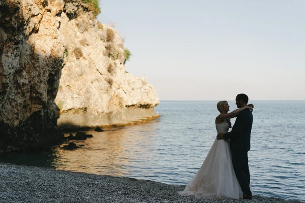 Groom and bride embrace beside a white limestone rock — calm sea and clear sky behind