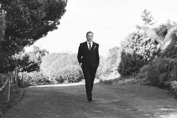 Black and white candid shot of the groom in a dark suit walking confidently along a garden path at the Royal Botanic Garden Edinburgh. Documentary portrait.