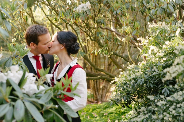 The couple share a kiss surrounded by white-flowering rhododendron bushes in the Royal Botanic Garden Edinburgh. Romantic documentary wedding portrait.