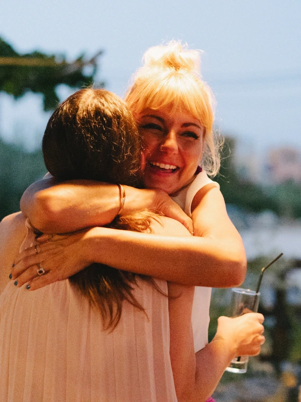 Two women embrace laughing outdoors in summer sunlight — blonde in pink dress, sea and green behind