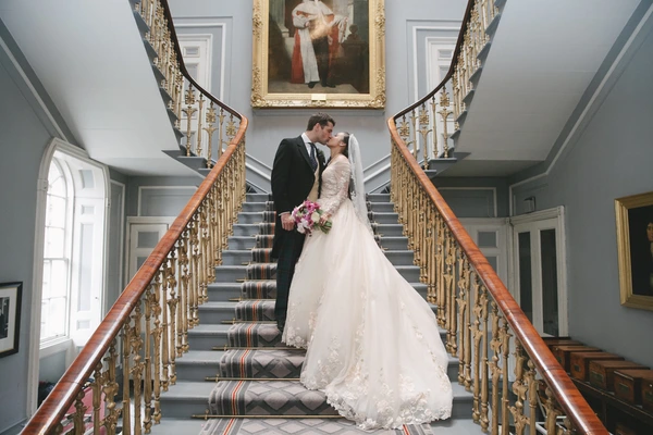 Newlyweds kiss on an elegant staircase — symmetrical gilded railings and painting on the wall