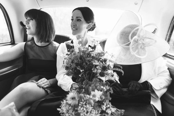 Black and white candid shot of the bride in her Norwegian bunad holding her bouquet in a taxi with her bridesmaid and mother, whose face is hidden by her large fascinator hat. Playful documentary moment.