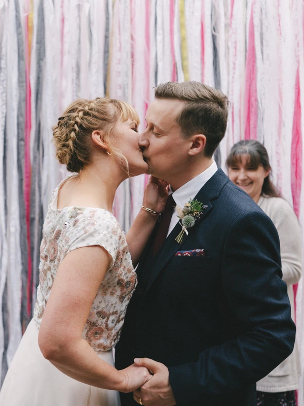 Bride and groom kiss in front of the striped backdrop, a happy guest watching behind