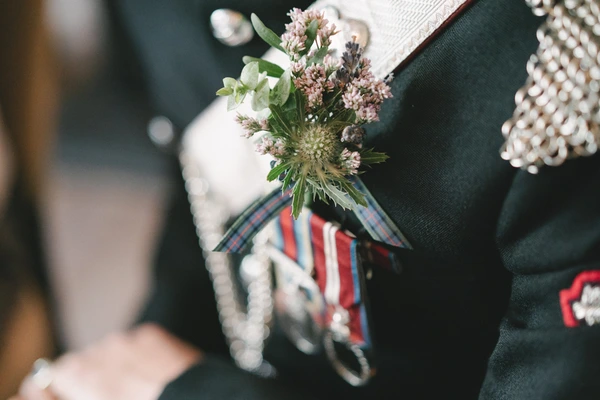 Groom in green suit holds the bridal bouquet — rust red and white flowers, tricolour ribbon