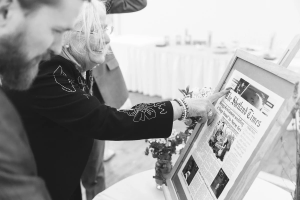 Black and white candid shot of two guests examining a framed copy of The Shetland Times newspaper featuring a wedding front page, laughing as they point at the article. Personalised wedding detail.
