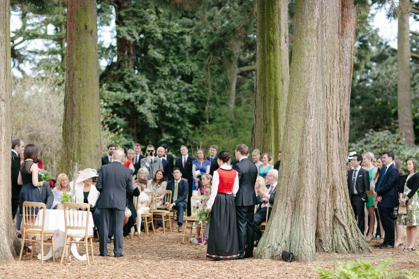 Wide colour shot of the outdoor ceremony from behind the couple, guests seated and standing under tall redwood trees at the Royal Botanic Garden Edinburgh. Documentary wedding scene.