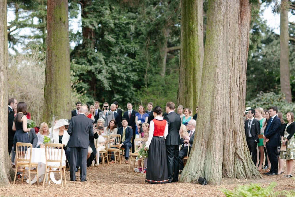 Wide colour shot of the outdoor ceremony from behind the couple, guests seated and standing under tall redwood trees at the Royal Botanic Garden Edinburgh. Documentary wedding scene.