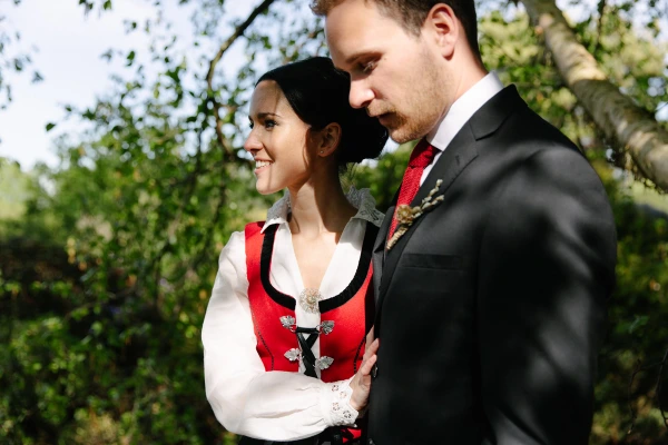 Bride in her Norwegian bunad smiling up at the groom in a dark suit and red tie while standing close together against green foliage at the Royal Botanic Garden Edinburgh. Romantic couple portrait.