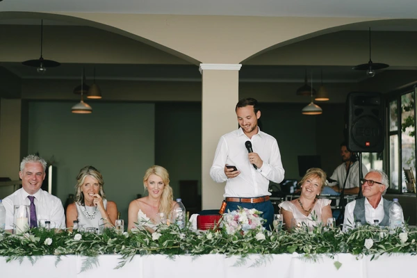 Groom speaks smiling with a microphone at the table, guests listening in a green-walled room