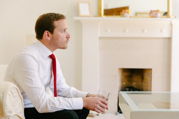 Groom in a white shirt and red knit tie sitting pensively by a fireplace, holding a glass of whisky. Quiet pre-ceremony moment.