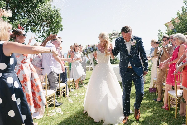Bride and groom walk the aisle while guests throw confetti — green trees, sunny garden