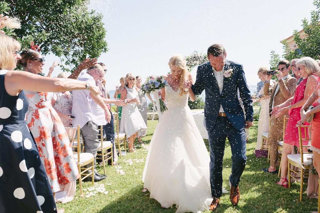 Bride and groom walk the aisle while guests throw confetti — green trees, sunny garden