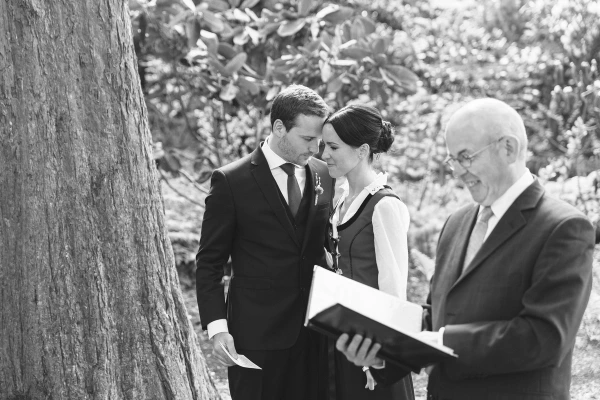 Groom looking intently at the bride while holding a vows card, the couple facing each other with the ceremony officiant visible to one side. Candid documentary moment.
