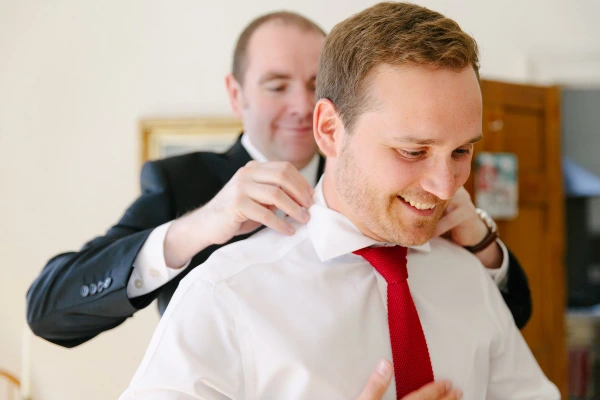 Groomsman adjusting the collar of the smiling groom in a white shirt and red tie during wedding preparations. Candid moment between friends.