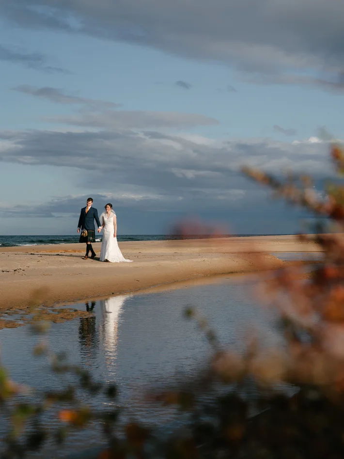 Bride and groom walk hand-in-hand on a golden Scottish beach, reflections shimmering in a tidal pool, dramatic cloudy sky behind them.