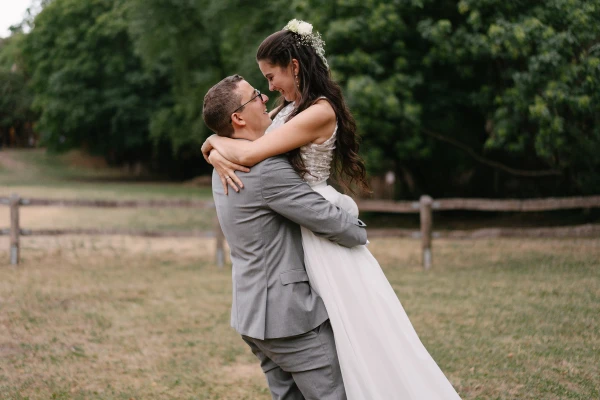 Groom lifting bride in white dress playfully at outdoor wedding with green trees and fence.