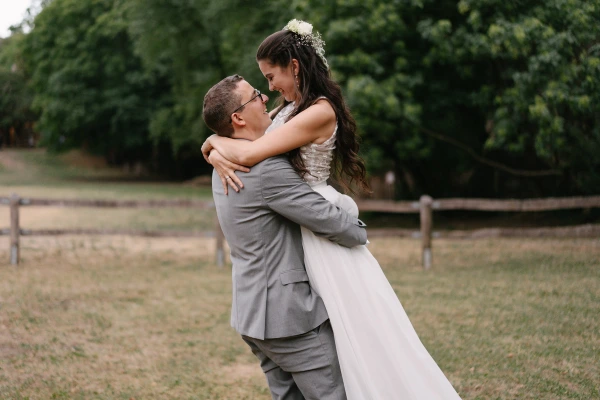 Groom lifting bride in white dress playfully at outdoor wedding with green trees and fence.