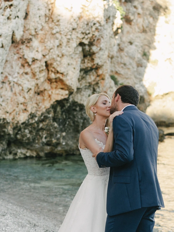 Bride and groom embrace beside sea rocks in front of turquoise water — intimate shore moment