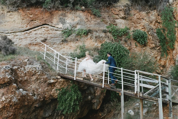 Bride and groom climb white metal steps on a rocky, plant-covered hillside — her dress flows gently