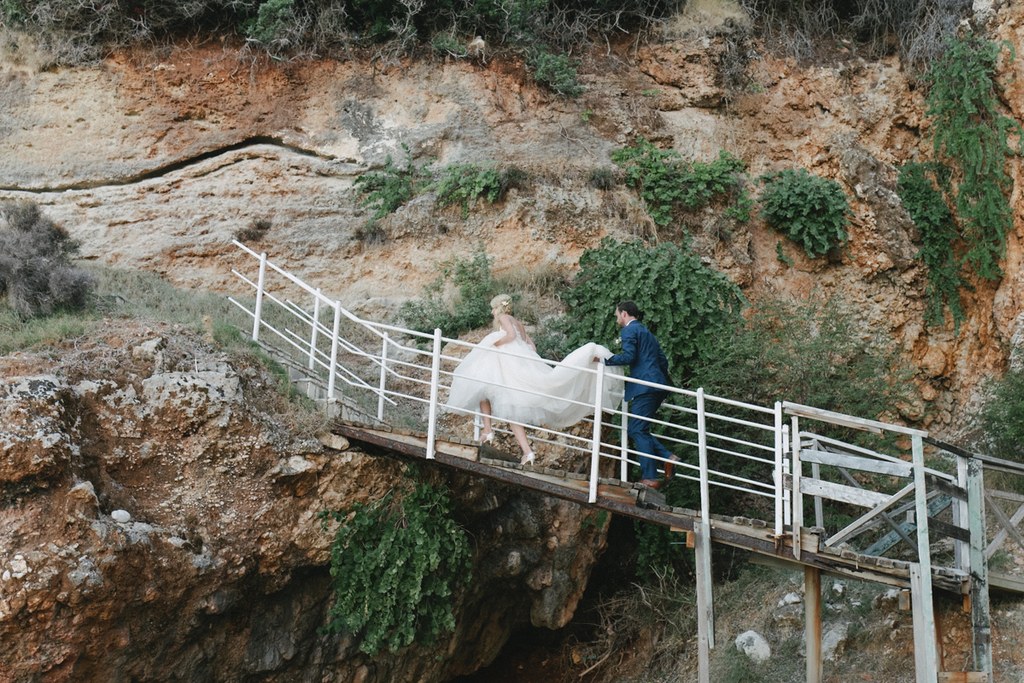 Bride and groom climb white metal steps on a rocky, plant-covered hillside — her dress flows gently