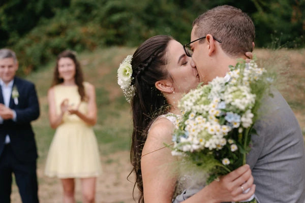 Bride and groom kissing during outdoor wedding ceremony with white daisy bouquet and guests in background.