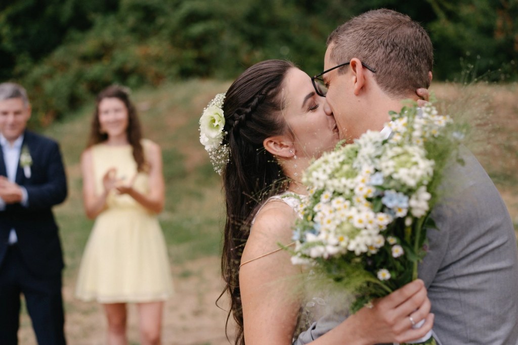 Bride and groom kissing during outdoor wedding ceremony with white daisy bouquet and guests in background.