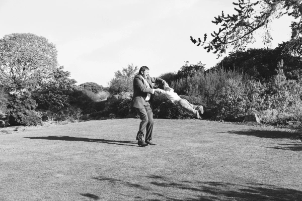Black and white image of a man in a suit spinning a child through the air on a garden lawn, both captured mid-movement. Candid and playful documentary wedding photography.