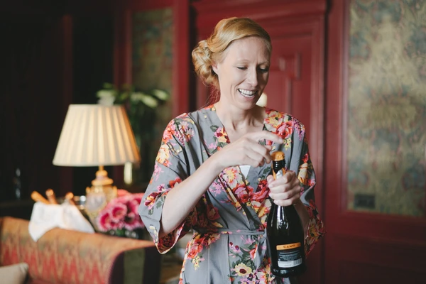 Red-haired woman in floral robe smiling while opening a champagne bottle in an elegant room