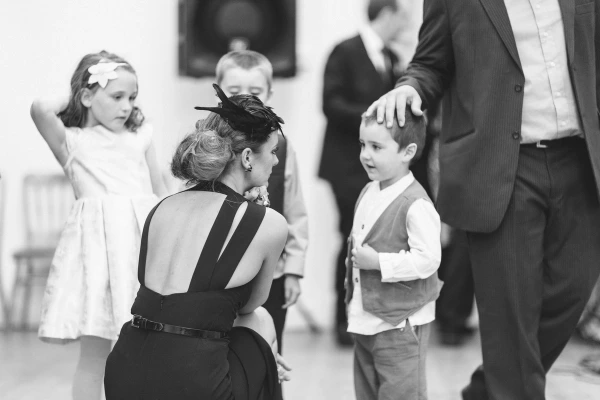 Black and white indoor shot of a guest in a black backless dress crouching to talk to a small boy in a waistcoat on the dance floor, with a flower girl watching nearby. Candid documentary wedding photography.