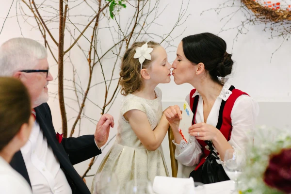 The bride in a Norwegian bunad and red vest exchanges a kiss with a flower girl in a cream dress at the wedding reception table. Candid and tender documentary moment.