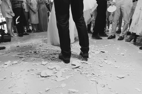 Groom's black shoes step among scattered flower petals, bride's white train beside him, guests around