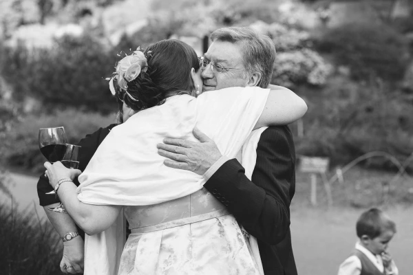 Black and white image of the bride leaning in to kiss an older man on the cheek in the garden while he holds a glass of red wine. Candid documentary wedding moment.