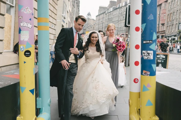 Groom leads bride by the arm through the city centre — long lace train, colourful pillars behind