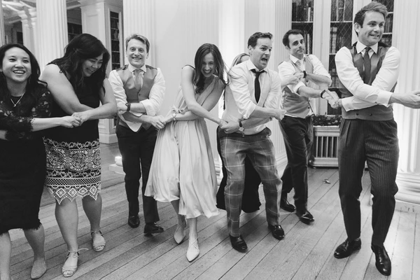 Wedding guests link arms and dance in an elegant colonnaded hall, big smiles all around