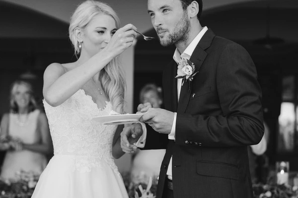 Bride with full mouth offers a spoon to the groom, he smiles — black-and-white, guests behind