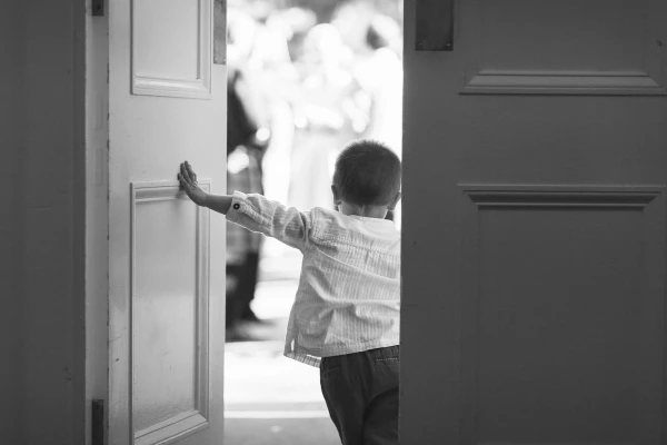 Black and white candid shot of a young boy pushing open a large door and peering out toward the bright garden where guests are gathered at the reception. Charming documentary moment.