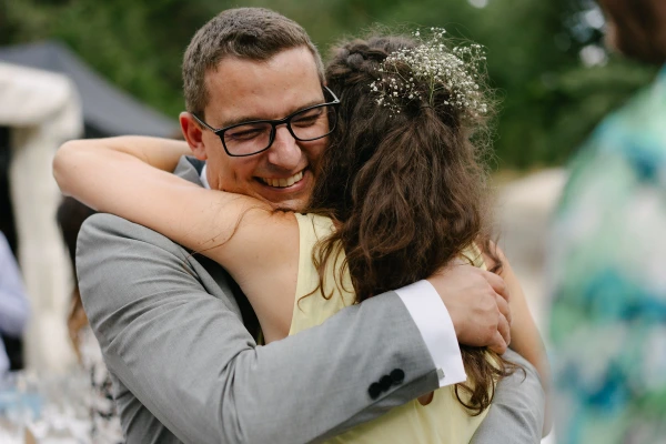 Groom hugging bridesmaid in yellow dress with baby's breath crown at outdoor wedding celebration.