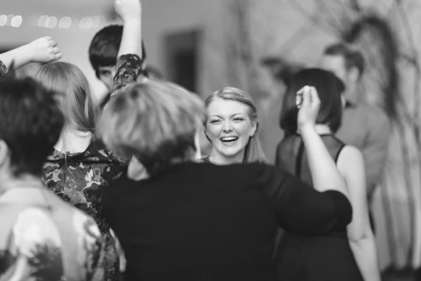 Black and white image of guests dancing energetically at the wedding reception, a young man mid-move and a flower girl being swung around in the background. Candid documentary moment.