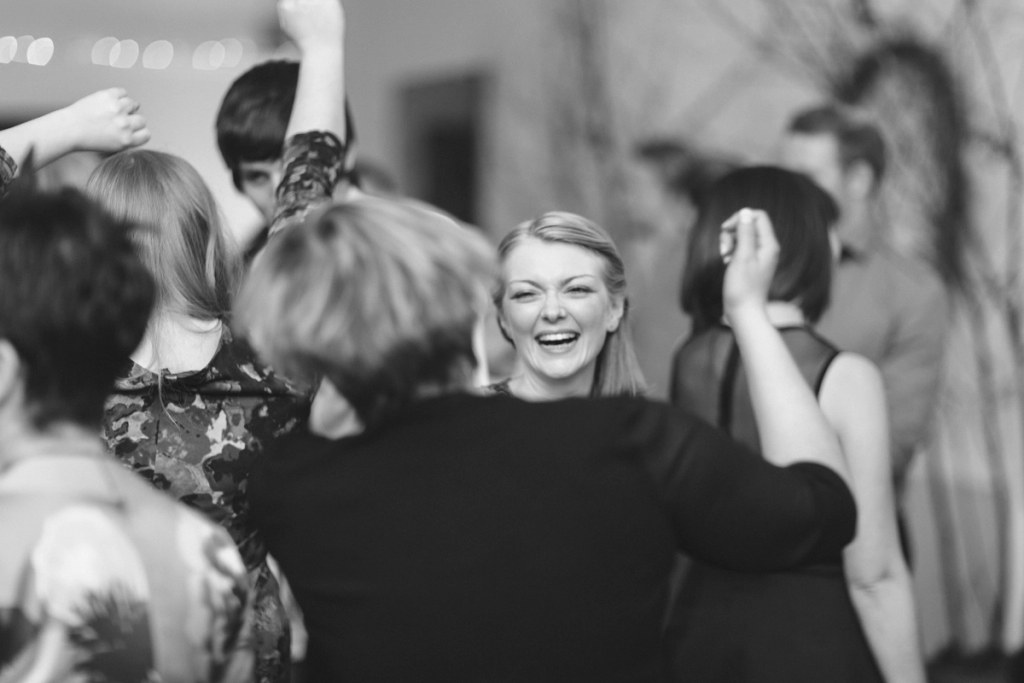 Black and white image of guests dancing energetically at the wedding reception, a young man mid-move and a flower girl being swung around in the background. Candid documentary moment.
