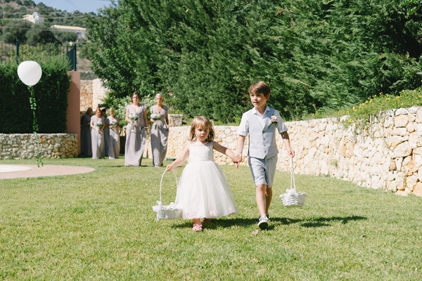 Two children walk hand in hand on a green lawn — girl with flower basket in white lace, boy in suit