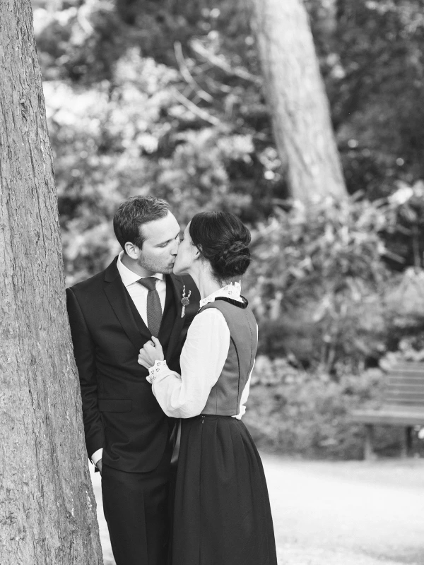 Black and white image of the groom leaning against a large tree to kiss the bride in her Norwegian bunad. Intimate documentary wedding portrait at the Royal Botanic Garden Edinburgh.