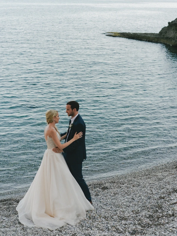 Newlyweds romantic moment on the beach — white sequined gown, dark suit, pebble shore, sea and rocky cliffs